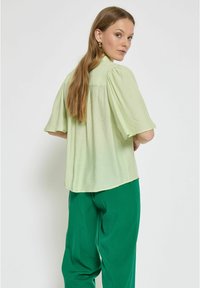 Blusa verde chiaro a maniche corte con vestibilità rilassata, tessuto texturizzato e uno scollo arricciato sul retro, abbinata a pantaloni verdi.
