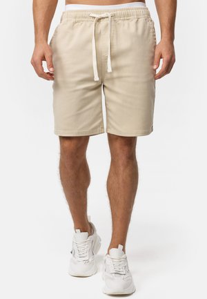 Beige cotton shorts with an elastic waistband and drawstring. Features side pockets and a straight leg design. Worn with white sneakers.