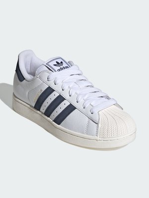 Biały sneaker Adidas Superstar z czarnymi paskami, konstrukcją z ochronnym czubkiem, logo na języku i kremową podeszwą, pokazany pod kątem na białym tle.
