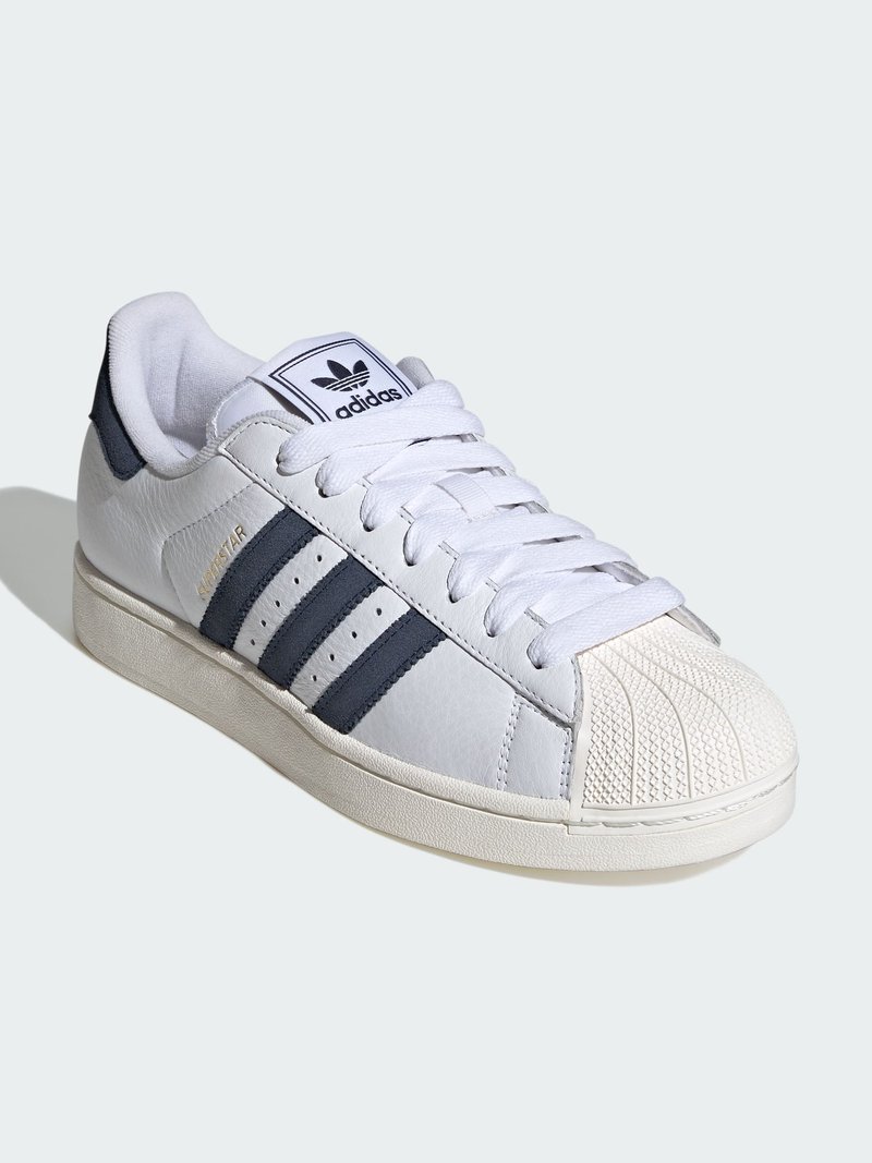 White Adidas Superstar sneaker with black stripes, shell-toe design, logo on tongue, and cream-colored sole, shown at an angle on a white background.
