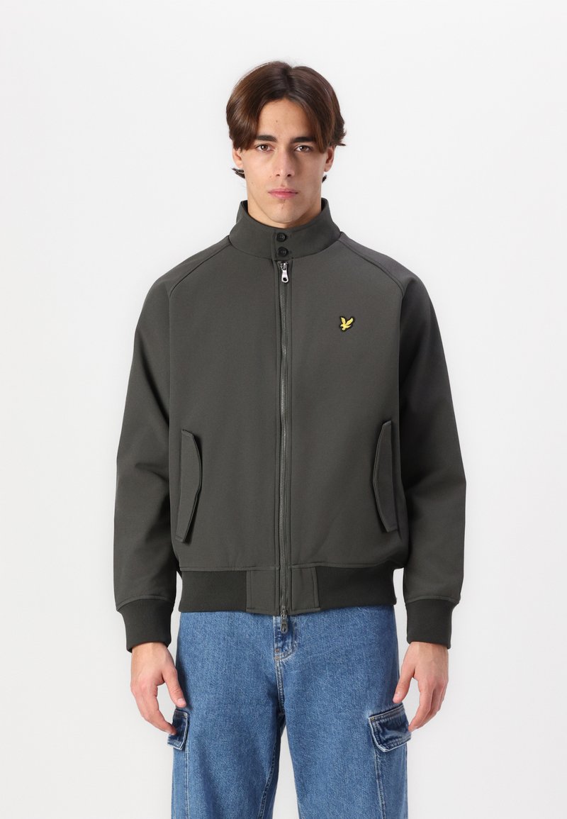 Giacca verde scuro con zip e colletto alto, tasche frontali, dettagli grigi e un piccolo logo giallo sul petto. Tessuto dalla superficie liscia.