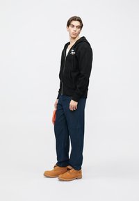 Μαύρο φούτερ με φερμουάρ και μπροστινές τσέπες, μπλε ριγέ παντελόνι και καφέ μπότες. Το μοντέλο κρατά μια πορτοκαλί τσάντα, παρουσιάζοντας ένα στρωμένο, καθημερινό ντύσιμο.