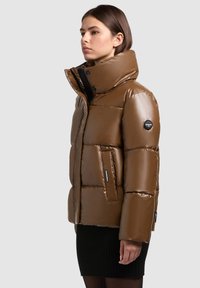 Jachetă puf din maroniu, cu guler înalt, textură lucioasă, închidere cu fermoar și un buzunar lateral. Prezintă un logo rotund pe braț.