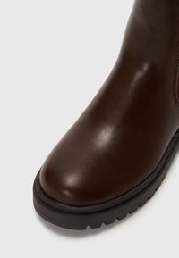 Bota de cuero marrón hasta el tobillo con puntera redondeada, acabado suave y suela texturizada. Presenta un sutil detalle de costura a lo largo del costado.