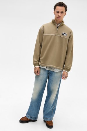 Ung mand iført en tan quarter-zip sweatshirt, en stribet skjorte nedenunder, slidte vide jeans og brune loafers, stående mod en hvid baggrund.