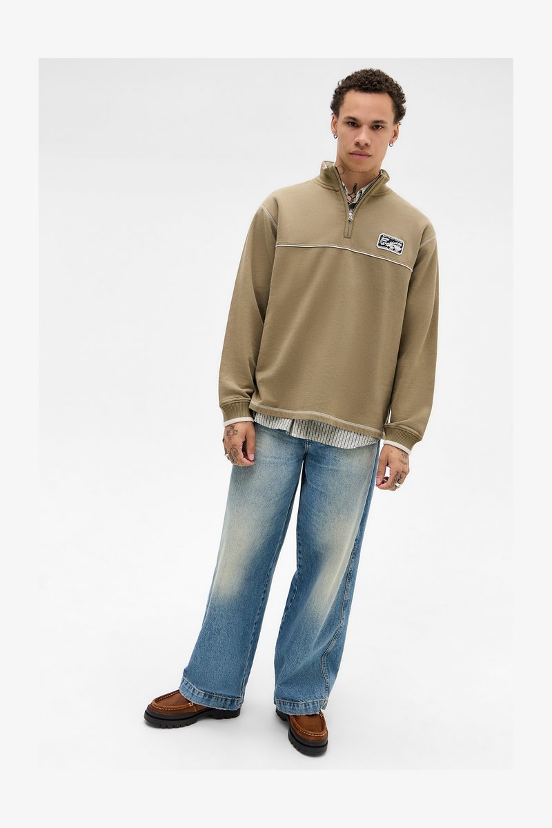 Jovem vestido com um suéter de pulôver bege de zíper até à metade, camisa listrada por baixo, calças largas desbotadas, mocassins castanhos, de pé contra um fundo branco.