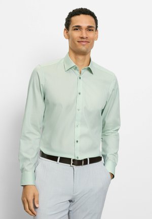 Mann trägt ein hellgrünes Hemd mit Knöpfen, hellgraue Hose und schwarzen Gürtel, steht mit einer Hand in der Tasche.