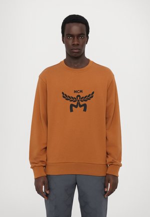 Brązowy sweatshirt z długimi rękawami, okrągłym dekoltem, z czarnym graficznym logo umieszczonym na środku klatki piersiowej. Miękka, bawełniana tkanina.