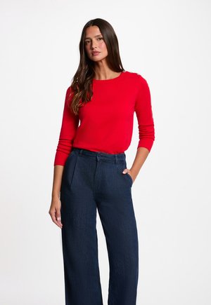 Mujer con el pelo largo y castaño, vestida con una blusa roja brillante de manga larga y pantalones anchos de talle alto azul oscuro, de pie con una mano en el bolsillo.