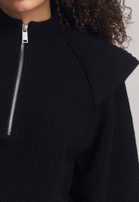 Pull en maille côtelée noire avec un col montant, des manches courtes structurées et une fermeture éclair argentée au niveau du col, mettant en avant un détail texturé.