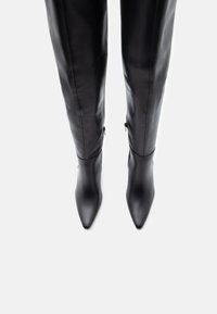 Ζευγάρι ψηλών μαύρων δερμάτινων μπότες με μυτερές μύτες και πλάγιες φερμουάρ, σε θέαση από επάνω σε λευκό φόντο.