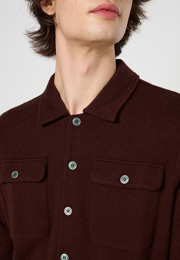 Jongere in een donkerbruine overhemd met knopen en twee borstzakken, gezien van nek tot middel, lichtjes naar links gericht.