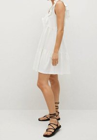 Weißes ärmelloses Kleid mit Rüschenträgern, geschichteter Gestaltung und leichtem Stoff. Getragen mit schwarzen Sandalen mit Riemen.