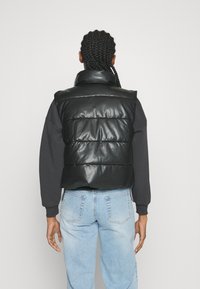 Svart vadderad väst i glänsande material, har en hög krage och en figurnära design, bärs över en långärmad svart skjorta och blå jeans.