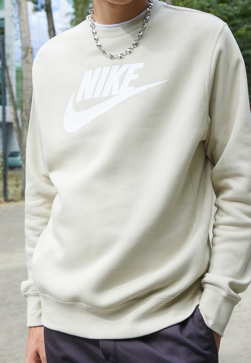 En person som bär en krämfärgad Nike-tröja och silverhalsband, står utomhus med träd och byggnader i bakgrunden.