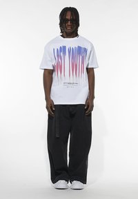 Baltas didelis T-shirt su mėlynu ir rožiniu lašelių dizainu "LOST YOUTH". Rūbą užbaigia juodos plačios kelnės ir baltos sportbačiai.
