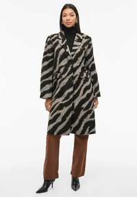 Kvinde iført en sort og grå zebra-mønstret frakke, sort turtleneck, brune bukser og sorte støvler med hæl, der står foran en hvid baggrund.