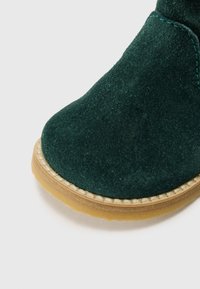 Botín de ante verde oscuro con punta redondeada y suela de goma marrón. Presenta costuras en contraste a lo largo del borde de la suela.