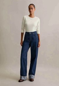 Mulher vestindo uma camisa branca de manga longa, jeans largos de cintura alta em azul escuro com barra, e sapatos marrons, em pé contra um fundo liso.