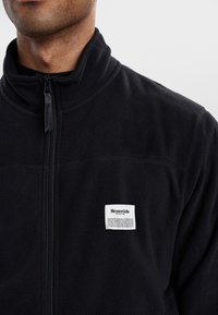 Giacca di pile nera con colletto alto, chiusura con zip e un'etichetta bianca del marchio sul lato sinistro del petto, caratterizzata da una texture morbida.