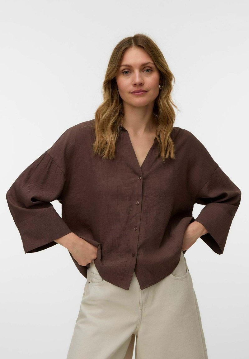 Femme aux cheveux ondulés châtain clair, portant une chemise marron ample à boutons et un pantalon beige, debout les mains dans les poches devant un fond uni.