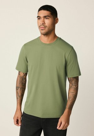 Lühikeste juustega mees, kes kannab lihtsat oliivrohelist T-särki ja mustasid pükse, seisab neutraalse tausta ees ning näitab mõlema käe tätoveeringuid.