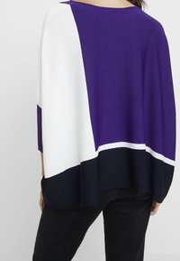 Violetti, valkoinen ja tummansininen väriestetty poncho, jossa on leveä kaula-aukko ja löysä istuvuus, valmistettu pehmeästä kankaasta. Korostuksena on kontrastiväriset reunat.