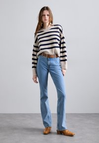 Gestreifter Strickpullover in Beige und Marineblau, blaue Schlagjeans und braune Lederschuhe mit runder Spitze. Ein einfacher Gürtel betont die Taille.