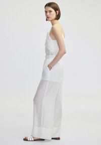 Weißer, ärmelloser Jumpsuit mit geschichteter, diagonalem Stoffdesign. Lockerer Schnitt, elastischer Bund, durchsichtige Abschnitte und integrierte Taschen.