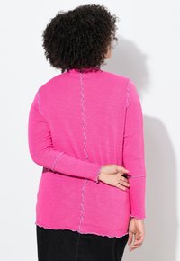 Vaaleanpunainen pitkähihaisen yläosa, jossa on aaltoileva reuna, nappimallinen kaulus ja kontrastiväriset vaaleansiniset saumat selässä ja hihoissa.