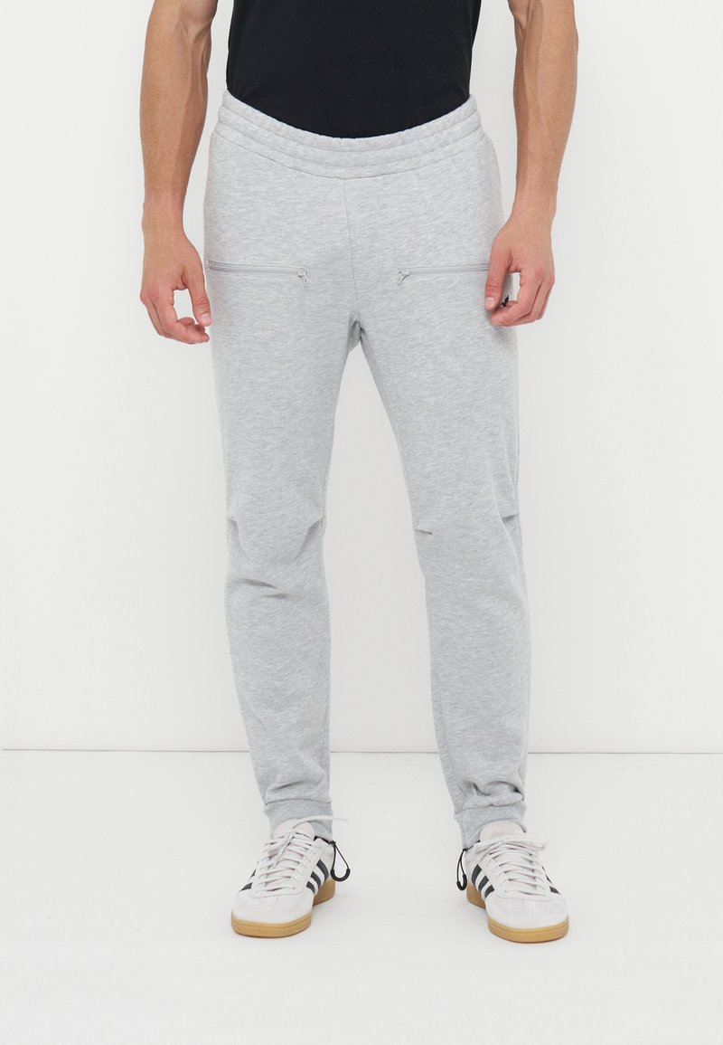 Άνδρας που φορά ανοικτό γκρι φόρμα με εμπρόσθιες τσέπες με φερμουάρ, λευκά και μαύρα αθλητικά παπούτσια, στέκεται μπροστά σε απλό λευκό φόντο.