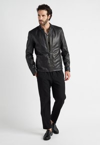 Zwarte leren jas met een opstaande kraag, geribbelde schouderdetails en een ritssluiting aan de voorkant. Gedragen over een gestreepte shirt en gecombineerd met zwarte broeken.