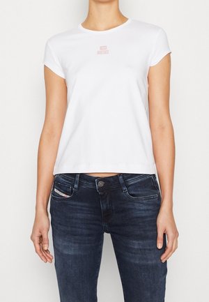 Vrouw draagt een nauwsluitend wit Diesel T-shirt en donkerblauwe jeans, staat met de armen ontspannen langs haar zij tegen een effen achtergrond.