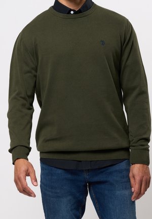 Man draagt een donkergroene trui met ronde hals over een zwart overhemd met kraag en blauwe spijkerbroek, staand met ontspannen armen naast het lichaam.