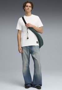 Weißes T-Shirt, lockere Passform, kombiniert mit hellblauen, ausgestellten Jeans. Eine grüne, gesteppten Umhängetasche mit schwarzem Schultergurt und Schlüsselanhänger-Detail.