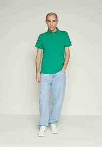 Camisa polo verde com colarinho, combinada com jeans de perna reta azul claro. A polo apresenta um pequeno logótipo no peito e mangas curtas.