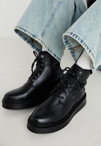 Must musta ankle saapad ümmarguste varvastega, nööridega disain ja paks must tald, koos helesiniste laia säärega teksapükstega.