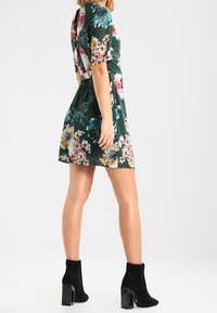Vestido floral en verde oscuro con mangas cortas, que presenta coloridas flores en rosa, blanco y azul. Botines negros hasta el tobillo con un tacón grueso.