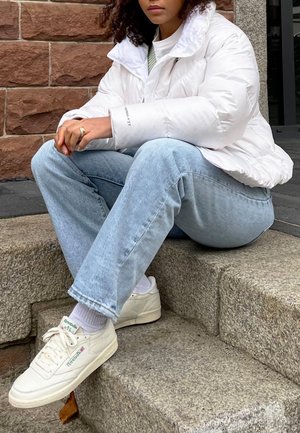 White puffer jacket with a high collar, light blue denim jeans, and white Reebok sneakers. Textured stone steps in the background.