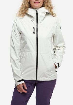 Femme portant une veste à capuche blanche avec une fermeture éclair noire et un pantalon violet, debout, une main dans la poche et l'autre détendue.