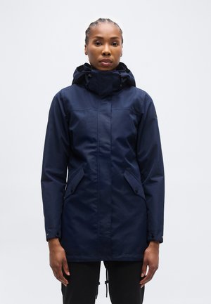 Žena oblečená v tmavomodrej nepremokavej bunde s kapucňou a vreckami s klopami na gombíky, stojaca pred jednoduchým svetlým pozadím.