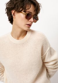 Beige gestrickter Pullover mit Rundhalsausschnitt und lockeren Ärmeln, kombiniert mit braun getönten Sonnenbrillen mit einem goldenen Rahmen.