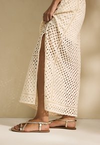 Vestito all'uncinetto color crema fino al ginocchio con motivo traforato e spacco laterale, abbinato a sandali piatti dorati con cinturino intrecciato e fibbia.