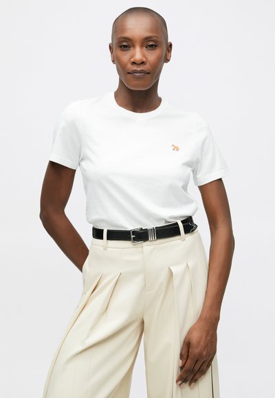 Bílý bavlněný tričko s malým oranžovým logem lišky na hrudi, spárované s elegantními béžovými kalhotami a černým opaskem.