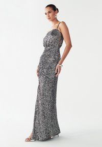 Fonkelende zilveren en zwarte avondjurk met een aansluitende pasvorm, dunne bandjes en een gedrapeerd voorpand. Voorzien van een zijsplit voor elegantie en bewegingsvrijheid.