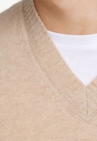 Beige gestrickter Pullover mit V-Ausschnitt und geripptem Design, über einem weißen Hemd getragen. Subtile Farb- und Weichkeitsunterschiede sind sichtbar.