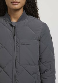 Grijze gewatteerde jas met een staande kraag, rits sluiting en een voorzak met logo. Materiaal heeft een gladde textuur en een lichtgewicht ontwerp.