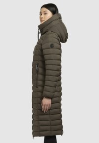 Long olive green puffer coat with a hood, featuring vertical quilted sections, side zip pockets, and a logo patch on the upper arm.