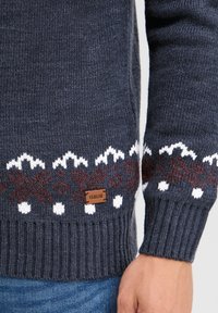 Gebreide trui in donkergrijs met witte en bordeauxrode sneeuwvlokpatronen, geribde manchetten en een klein bruin stoffen label met daarop "ISOLID."