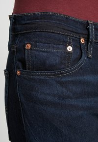 Primo piano di un paio di jeans di denim blu scuro che mostrano la tasca frontale con rivetti di rame e cuciture dettagliate, indossati con un top borgogna.